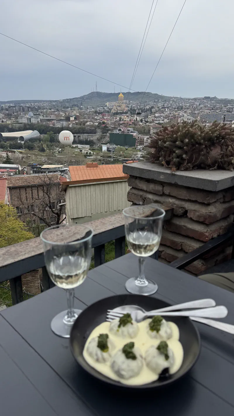Khinkali in a creamy sauce with two glasses of white wine on a terrace table above the Tbilisi skyline.